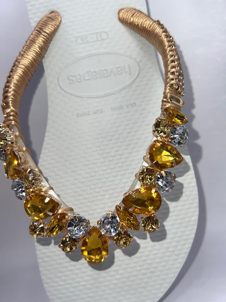 K&ouml;nnte beinhalten: Wei&szlig;e Havaianas-Zehensandale mit goldfarbenem Riemen, verziert mit goldenen, gelben und klaren Edelsteinen. Der Riemen ist mit goldfarbenem Material umwickelt. Die Zehensandale ist Gr&ouml;&szlig;e 37-38, was der EU-Gr&ouml;&szlig;e 39/40 entspricht.