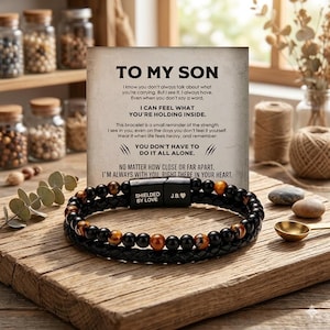 May include: A black and brown beaded bracelet with a leather band, featuring the text "SHIELDED BY LOVE" and initials "J.B.". A card with the text "TO MY SON" is behind the bracelet. The bracelet is on a wooden surface.