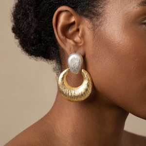 May include: A two-tone drop earring featuring a textured silver-colored oval stud at the top connected to a large, textured gold-colored crescent-shaped hoop. The metal surfaces have a brushed, irregular finish that catches the light.
