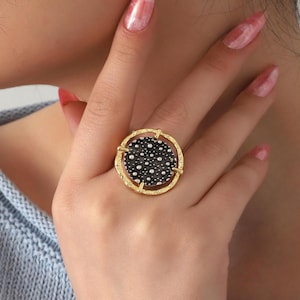 Op de afbeelding: Een goudkleurige ring met een grote, cirkelvormige voorkant. De voorkant heeft een zwarte achtergrond met talrijke kleine, witte stippen. De band van de ring heeft een textuurontwerp en de voorkant wordt omlijst door een gouden rand met kruisachtige accenten.