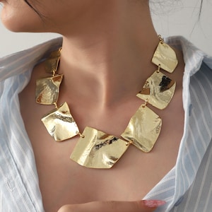 May include: A gold-coloured statement necklace featuring irregularly shaped, textured metal pieces. The necklace is composed of several connected, abstract geometric shapes, creating a bold and modern design. The jewellery is worn over a light blue striped shirt.