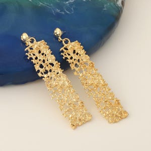 May include: A pair of gold-coloured, textured dangle earrings. Each earring has a small, round stud and a long, rectangular drop with an intricate, porous design. The earrings are displayed on a blue and white surface.