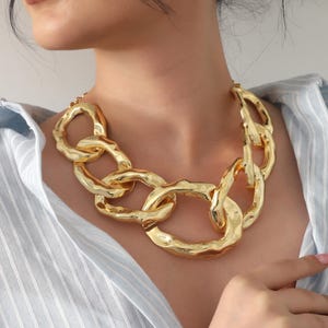 Puede incluir: Un collar grueso de tono dorado que presenta grandes eslabones ovalados interconectados con una superficie texturizada y martillada. El collar descansa alrededor del cuello, mostrando un acabado metálico audaz con formas orgánicas irregulares.
