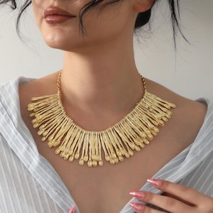 Può includere: Una collana importante color oro caratterizzata da una serie di elementi metallici verticali, testurizzati e a forma di cucchiaio, disposti a frangia lungo una catena. La collana poggia sulla clavicola, mostrando una finitura metallica uniforme.