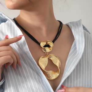 Könnte beinhalten: Eine goldfarbene Anhängerkette mit zwei miteinander verbundenen, texturierten, abstrakten, bandartigen Formen, die an einer schwarzen, mehrsträngigen Kordel hängen. Die Metalloberflächen haben ein gehämmertes, unebenes Finish, das das Licht einfängt und einen deutlichen Kontrast zur dunklen Kordel bildet.
