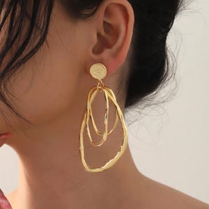May include: Gold-toned statement earrings. Each earring features a textured, circular stud that connects to three overlapping, irregular oval shapes. The earrings have a polished finish and are designed to dangle. The jewelry is a bold accessory.