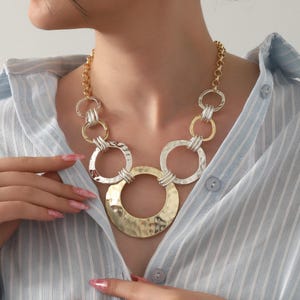 May include: A statement necklace featuring a mix of gold and silver tones. The necklace has a chain and a series of interconnected rings, with a large, hammered gold-tone pendant at the center. The jewelry is worn over a light blue striped shirt.