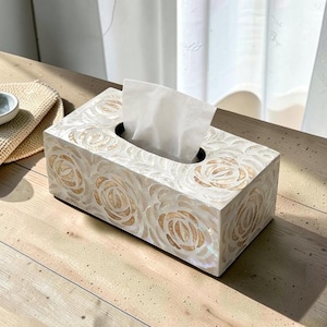 May include: Rectangular tissue box with a mother-of-pearl finish and a floral rose design. The box is white with gold accents and a black opening for tissue dispensing. A stack of white tissues is visible.