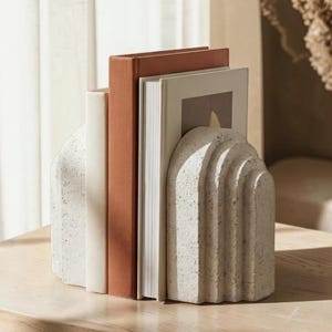 May include: A pair of textured, off-white bookends with an arched design, holding several books. The books have covers in shades of white, terracotta, and beige. The bookends and books are on a light-colored wooden surface.