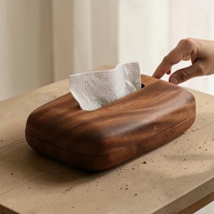 May include: A wooden tissue box with a smooth, curved design. The box is a rich brown color, and a white tissue is partially pulled out of the top opening. The tissue box is sitting on a light-colored wooden surface.