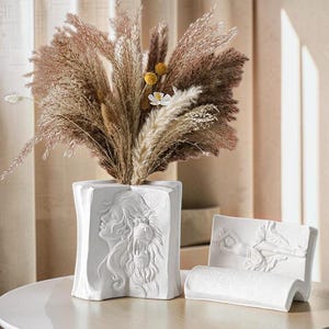 Peut inclure: Un vase en céramique blanche en forme de livre ouvert avec un bas-relief représentant une personne et un lion, contenant des herbes de la pampa séchées. À côté se trouve un objet en céramique blanche en forme de livre avec un bas-relief de personnage.