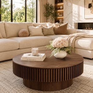 May include: A round, dark wood coffee table with a slatted base design. On the tabletop sits a white ceramic vase filled with light pink hydrangea flowers, a clear glass candle, and two stacked books titled 100 and AUTHENTICS.