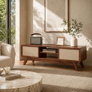 May include: A mid-century modern style wooden media console with a light-colored woven fabric panel on the doors. A small speaker, framed art, and a vase with greenery sit on top. The console has a shelf with books and is set on angled legs.