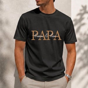 Op de afbeelding: Zwart T-shirt met ronde hals met het woord "PAPA" in een goudkleurig script. De woorden "Sempre & Can" staan eronder in een vergelijkbaar lettertype. Het shirt is gemaakt van een zacht materiaal.