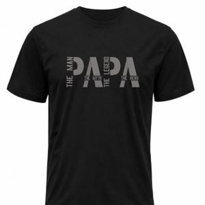 Op de afbeelding: Zwart T-shirt met ronde hals en het woord "PAPA" in grijs, met de zinnen "THE MAN", "THE MYTH", "THE LEGEND" en "THE HERO" verticaal afgedrukt aan weerszijden van het woord.