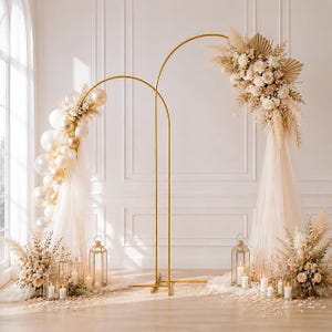 May include: Two gold metal arched frames decorated with white and gold balloons, beige pampas grass, dried palm leaves, and white flowers. Sheer white fabric drapes from the arches, with gold metal lanterns and floral arrangements on the floor.