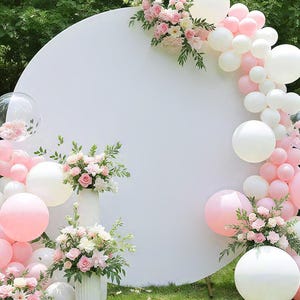 May include: A large white circular backdrop decorated with a balloon garland in shades of white and soft pink, accented with arrangements of pink and white roses and green foliage, featuring two white fluted vases filled with floral bouquets.