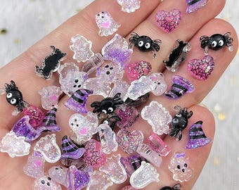 Könnte beinhalten: Eine Sammlung kleiner, dekorativer Halloween-Harz-Cabochons mit glitzernden Geistern, schwarzen Spinnen mit weißen Augen, violett-schwarz gestreiften Hexenhüten und rosa Herzen mit schwarzen Spinnennetzmustern in verschiedenen Größen.