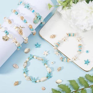 May include: A collection of stretch bracelets featuring turquoise blue starfish charms, white shell beads, and gold-toned accents. The set includes multiple bracelets displayed on a white cylindrical stand and a white rectangular tray with loose beads.