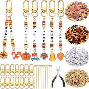 May include: A DIY keychain making kit featuring gold lobster clasps, metal pins, pliers, and assorted beads. Includes white letter beads spelling names like John, Emily, and David, plus autumn-themed charms like pumpkins, foxes, and leaves.