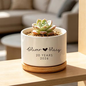 May include: A white ceramic cylindrical planter with a bamboo saucer base containing a small green succulent. The planter features black printed text that reads Oliver, a heart symbol, Stacy, and the text 20 YEARS 2026 below the names.