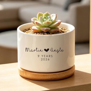 Op de afbeelding: Een witte keramische plantenpot met een vetplant, op een houten basis. De pot heeft de tekst "Martin ❤ Lingle" en "9 YEARS 2026" in zwart schrift. De vetplant heeft groene en rode bladeren.