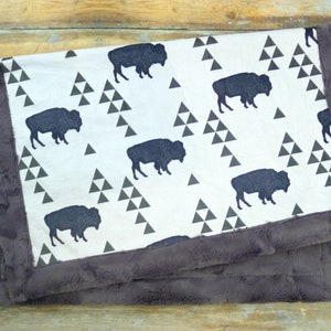 Puede incluir: Una manta de bebé blanca con un borde de piel sintética marrón. La manta presenta un patrón repetido de siluetas de búfalos negros y triángulos grises.