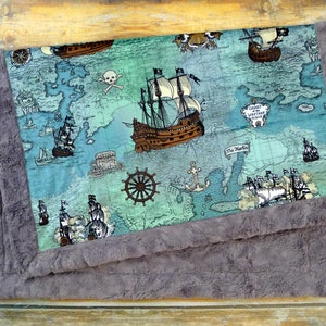 May include: A baby blanket featuring a pirate map design with ships, a skull and crossbones, and the text "Chart of the Western Ocean". The blanket has a soft, gray, plush border.