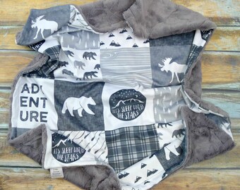 May include: A gray and white patchwork baby blanket with a plush gray backing. The blanket features a variety of woodland animal prints, including moose, bears, and trees. The blanket also includes the text "Let's sleep under the stars" and "Adventure".