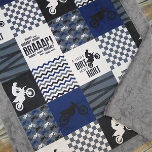 May include: A blue and gray baby blanket with a patchwork design featuring motocross bikes, checkered patterns, and the text "A little dirt never hurt" and "BRAAAP!"