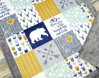May include: A baby blanket with a patchwork design featuring a bear, deer, arrows, mountains, and the text "I love you to the mountains and back". The blanket is made of soft, plush fabric and has a gray border.