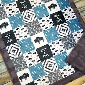 May include: A patchwork baby blanket with a blue, white, and gray color scheme. The blanket features a repeating pattern of buffalo silhouettes, geometric designs, and the text "Wild & Free".
