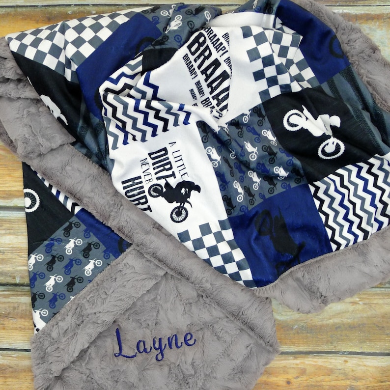 May include: A gray and blue patchwork baby blanket with a dirt bike design and the text "A Little Dirt Never Hurt". The blanket has a gray faux fur border and is personalized with the name "Layne".