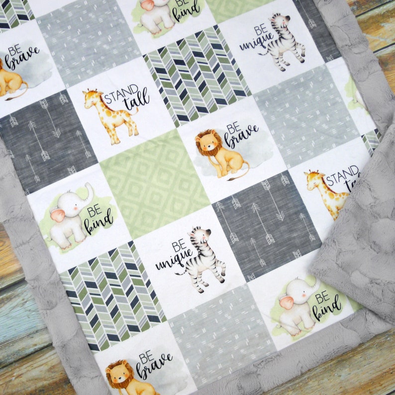 May include: A patchwork baby blanket with animal illustrations and motivational phrases. The blanket has a grey, textured border and features a giraffe, lion, elephant, and zebra. Phrases include "Be Brave", "Be Kind", and "Be Unique".