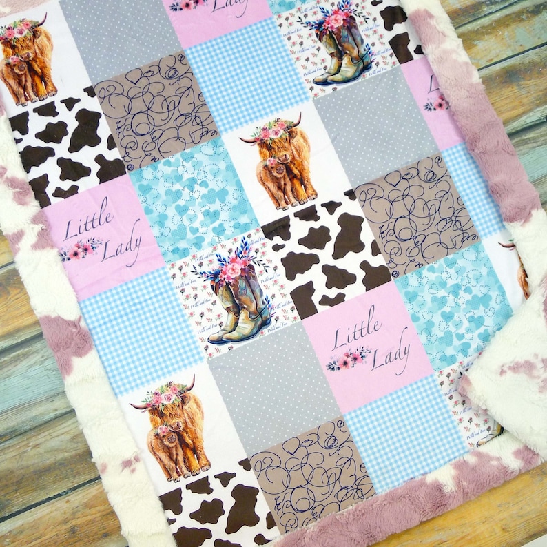 May include: A patchwork baby blanket featuring squares with cowhide patterns, Highland cows, and cowboy boots. The blanket includes the text "Little Lady" and is trimmed with a pink and white faux fur border.
