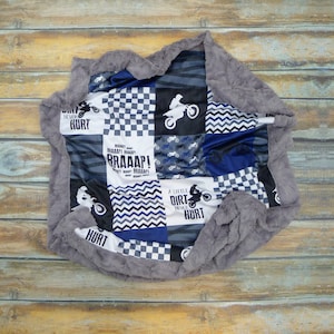May include: A gray and white patchwork baby blanket with a motocross theme. The blanket features a checkered pattern, a chevron pattern, and a pattern of dirt bikes. The blanket also has the text "A little dirt never hurt" and "BRAAAP!" printed on it.