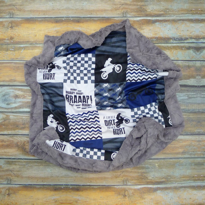 May include: A gray and white patchwork baby blanket with a motocross theme. The blanket features a checkered pattern, a chevron pattern, and a pattern of dirt bikes. The blanket also has the text "A little dirt never hurt" and "BRAAAP!" printed on it.