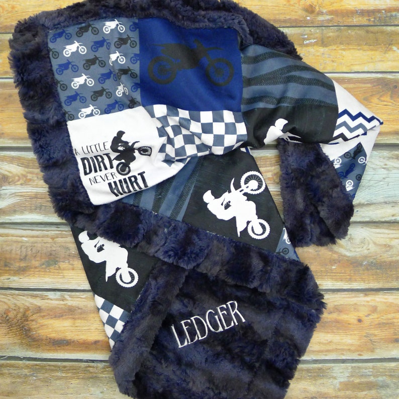May include: A blue and black patchwork baby blanket with a motocross theme. The blanket features a checkered pattern, a dirt bike silhouette, and the text "A little dirt never hurt". The blanket is personalized with the name "Ledger" embroidered in white on the bottom right corner.