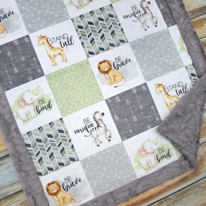 May include: A baby quilt with a grey, green, and white safari animal theme. The quilt features squares with illustrations of a giraffe, lion, elephant, and zebra, along with the phrases "BE BRAVE", "BE UNIQUE", "BE KIND", and "STAND TALL". The quilt has a grey, plush border.