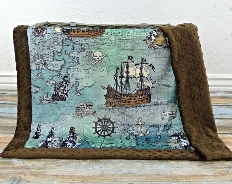 May include: A brown and blue pirate-themed blanket with a map design featuring ships, a skull and crossbones, and a ship's wheel. The blanket has a brown, textured border.
