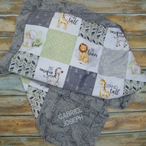 May include: A baby blanket in grey and white with a patchwork design. It features squares with animal illustrations, arrows, and the phrases "BE KIND", "BE BRAVE", "BE UNIQUE", and "STAND TALL". The blanket has a soft, plush border and the name "GABRIEL JOSEPH" embroidered on it.