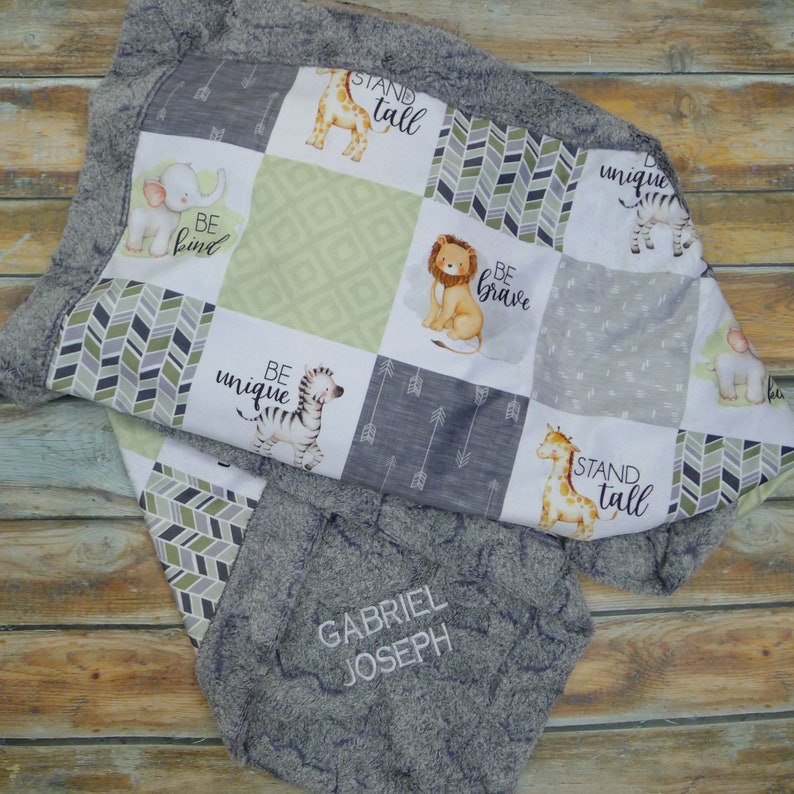 May include: A baby blanket in grey and white with a patchwork design. It features squares with animal illustrations, arrows, and the phrases "BE KIND", "BE BRAVE", "BE UNIQUE", and "STAND TALL". The blanket has a soft, plush border and the name "GABRIEL JOSEPH" embroidered on it.