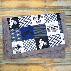 May include: A patchwork baby blanket with a gray fleece backing. The front of the blanket features a variety of designs including a checkered pattern, a chevron pattern, a dirt bike, and the text "A Little Dirt Never Hurt" and "BRAAAP!"