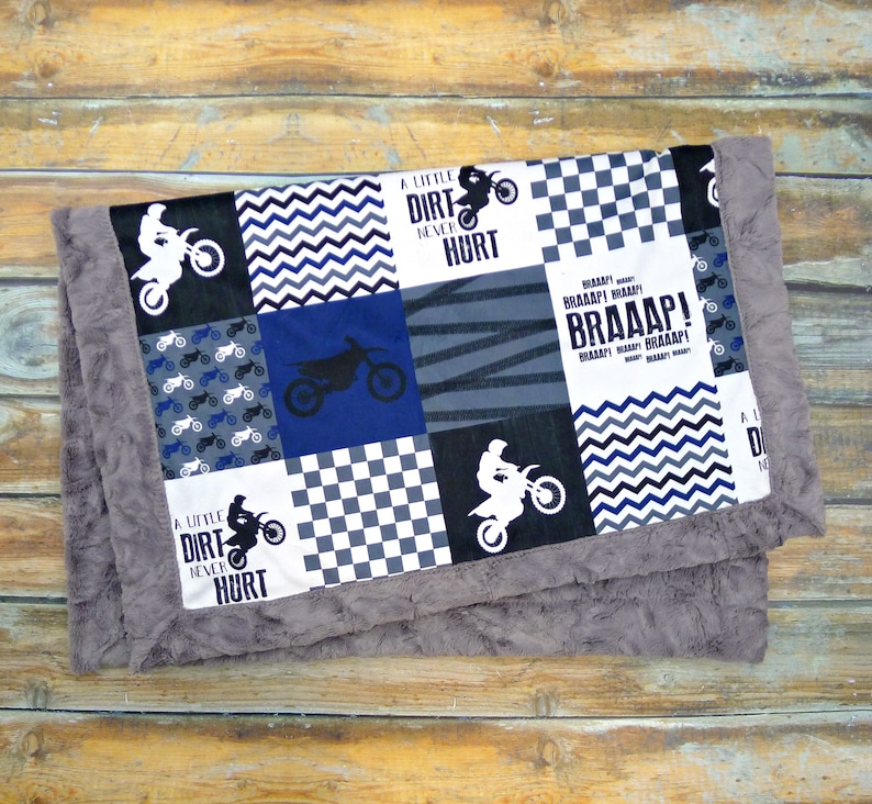 May include: A patchwork baby blanket with a gray fleece backing. The front of the blanket features a variety of designs including a checkered pattern, a chevron pattern, a dirt bike, and the text "A Little Dirt Never Hurt" and "BRAAAP!"