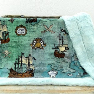 May include: A plush baby blanket with a pirate map design. The blanket features illustrations of pirate ships, crossed swords, and a skull and crossbones on a teal background. The edges are trimmed with a soft, light blue faux fur.