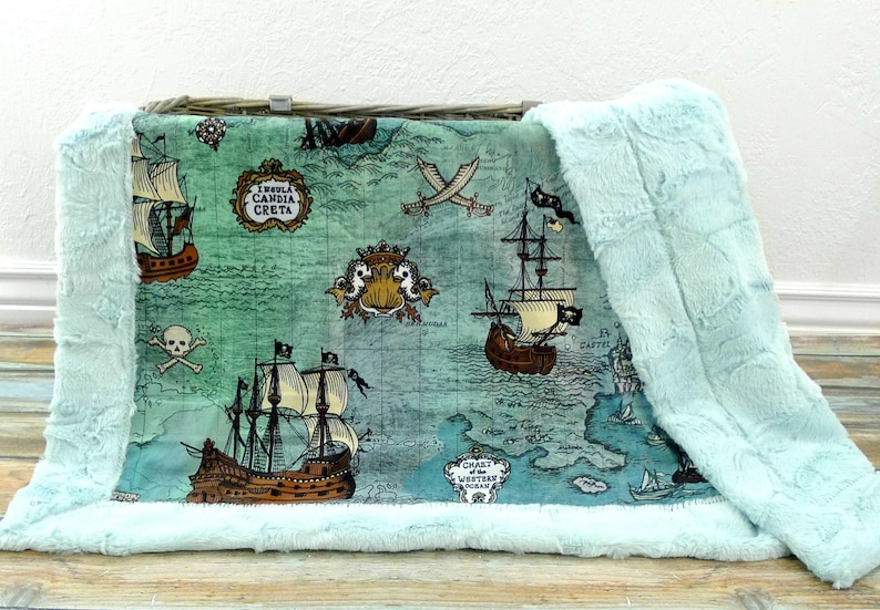 May include: A plush baby blanket with a pirate map design. The blanket features illustrations of pirate ships, crossed swords, and a skull and crossbones on a teal background. The edges are trimmed with a soft, light blue faux fur.