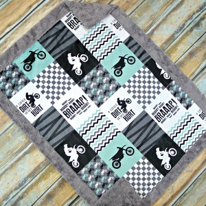 May include: A baby blanket with a patchwork design featuring a motocross theme. The blanket is made of soft, plush fabric and has a gray border. The blanket features a variety of designs, including a checkered pattern, a zebra print, and a chevron pattern. The blanket also features the words "A Little Dirt Never Hurt" and "BRAAAP!" in black text.