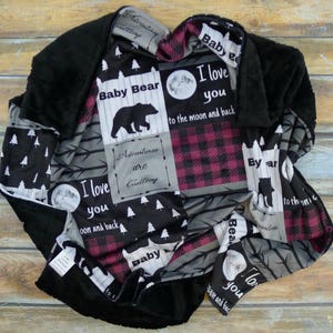 以下が含まれることがあります： クマ、木、そして「Baby Bear」、「I love you to the moon and back」、「Adventures are calling」というテキストが描かれたパッチワークデザインの黒と赤のベビーブランケットです。