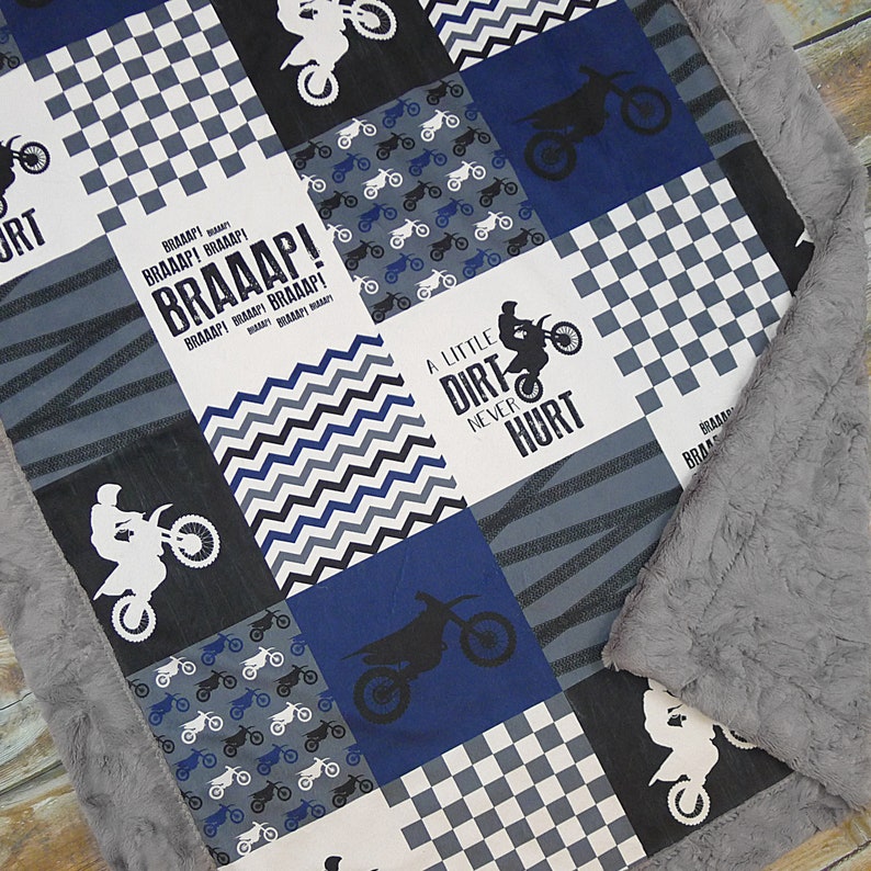 May include: A blue and gray baby blanket with a patchwork design featuring motocross bikes, checkered patterns, and the text "A little dirt never hurt" and "BRAAAP!"