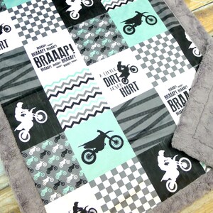 May include: A baby blanket with a patchwork design featuring a motocross theme. The blanket is made of soft, plush fabric and features a variety of colors, including gray, black, and teal. The blanket has a checkered pattern, a chevron pattern, and a pattern of dirt bike silhouettes. The blanket also features the text "A Little Dirt Never Hurt" and "BRAAAP!"